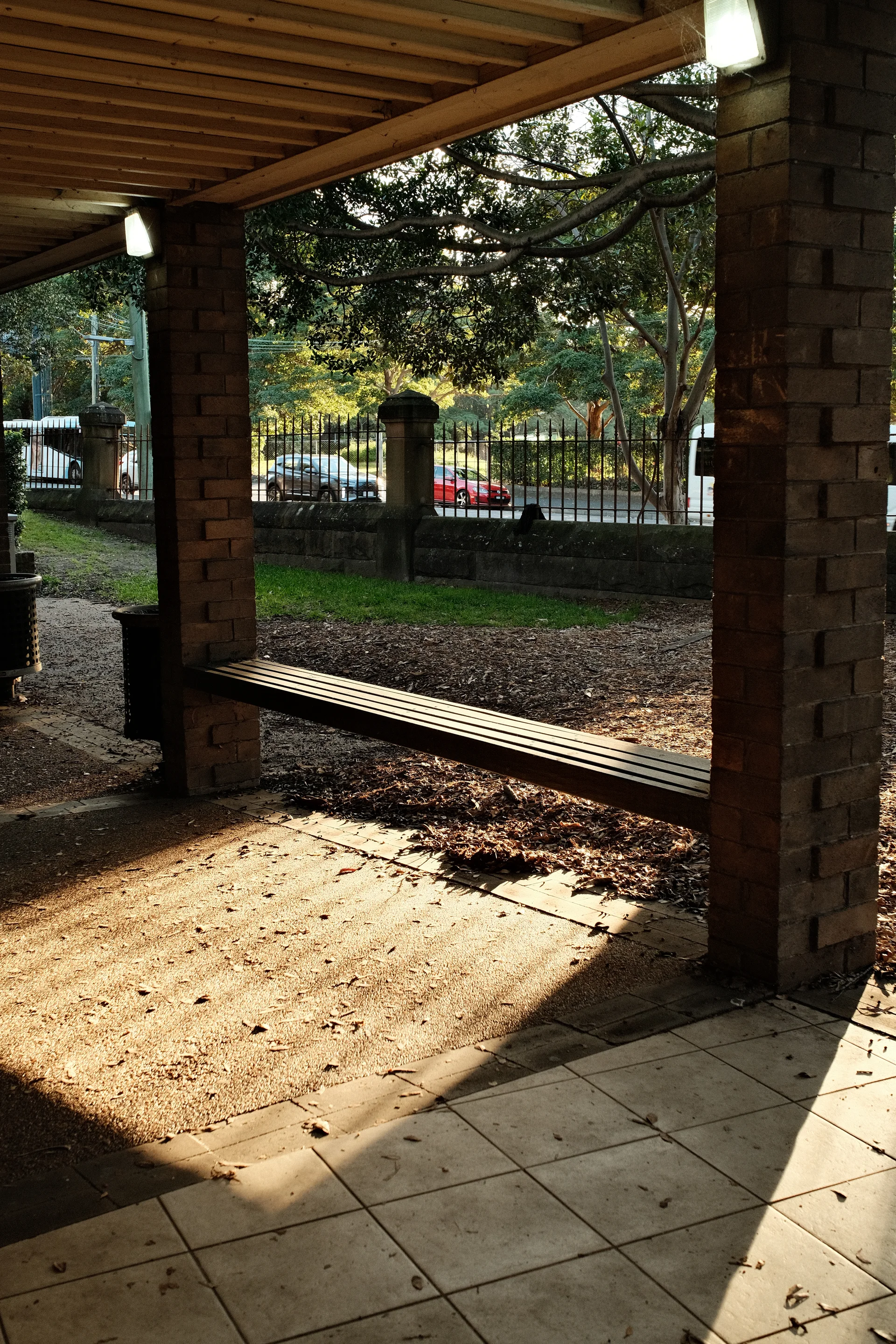 Sunlight crossing an empty timber bench beneath a brick shelter.