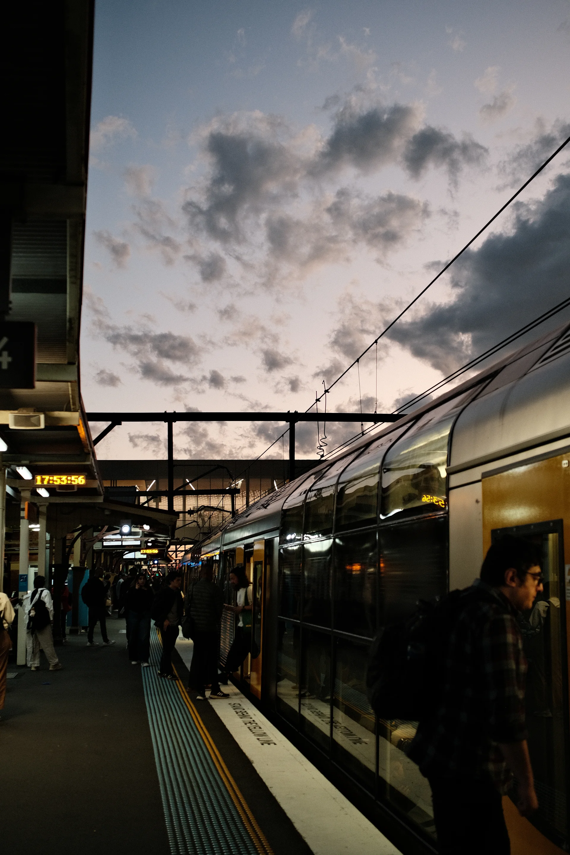 Commuters waiting beside a silver train beneath a dramatic evening sky.