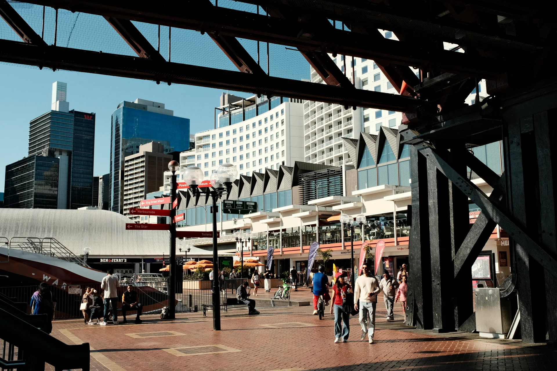 A pedestrian crossing a sunlit plaza beneath dark steel railway beams.