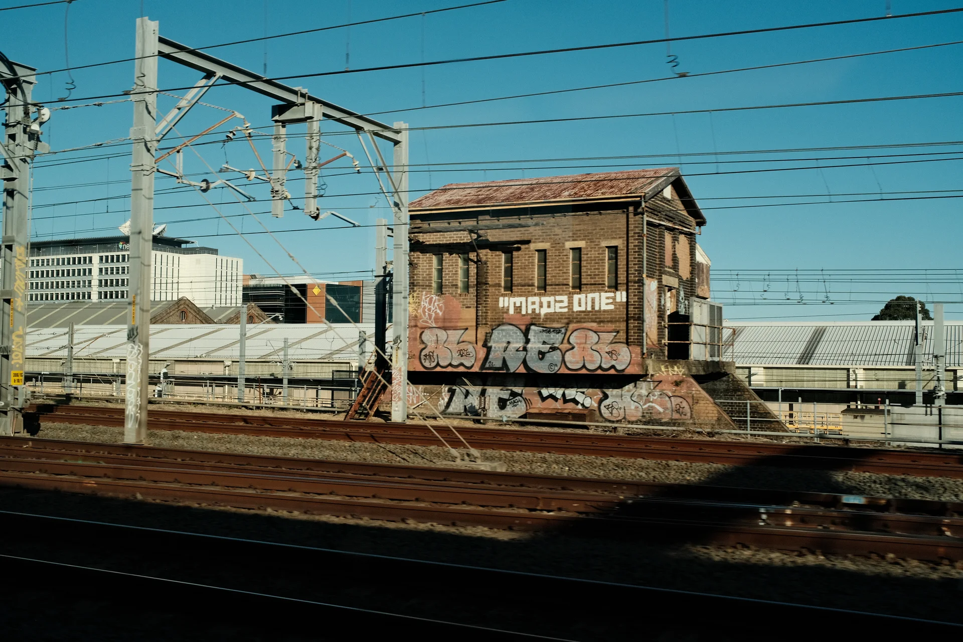 A graffitied brick railway building seen across tracks and overhead power lines.